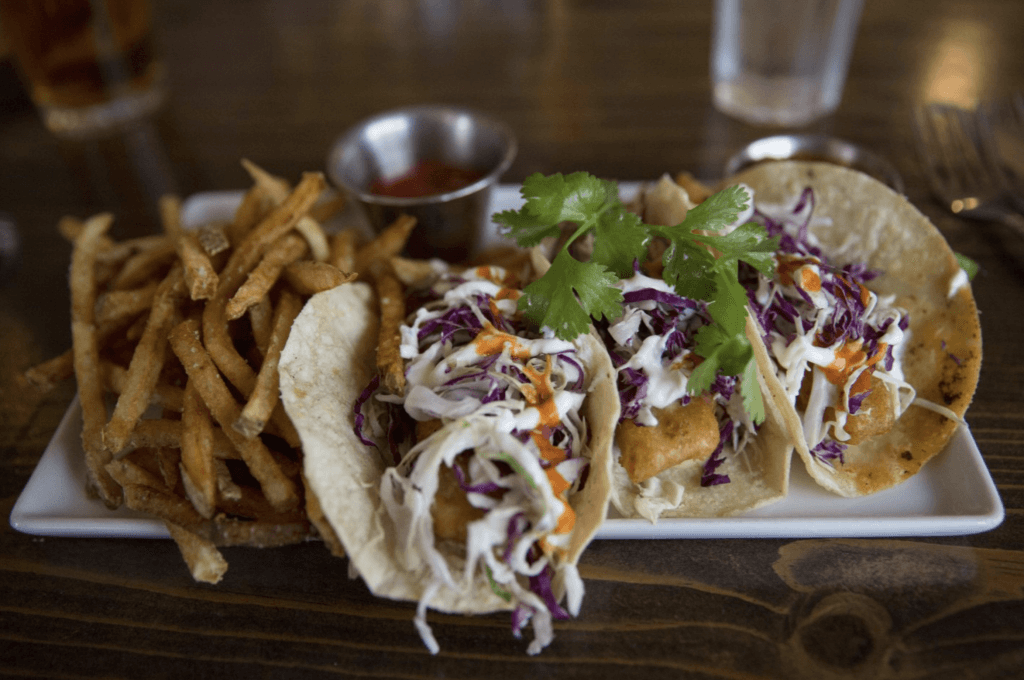 A close-up view of a plate of fries, a small cup of ketchup and three baja style fish tacos.